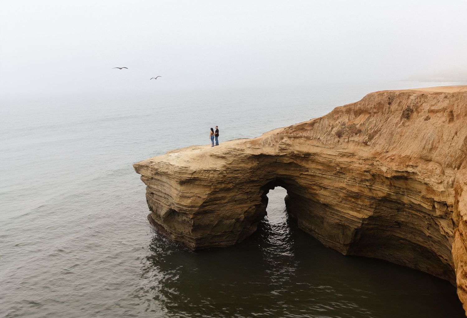 Two silhouettes stand on dramatic natural stone arch over ocean.