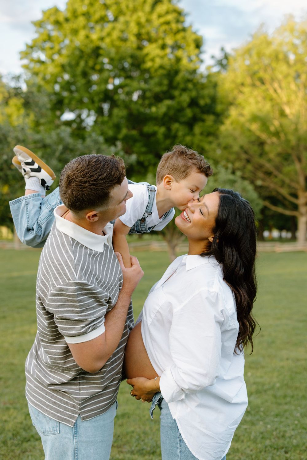 Happy family in park, father holding toddler son who kisses pregnant mother, sunny outdoor setting.