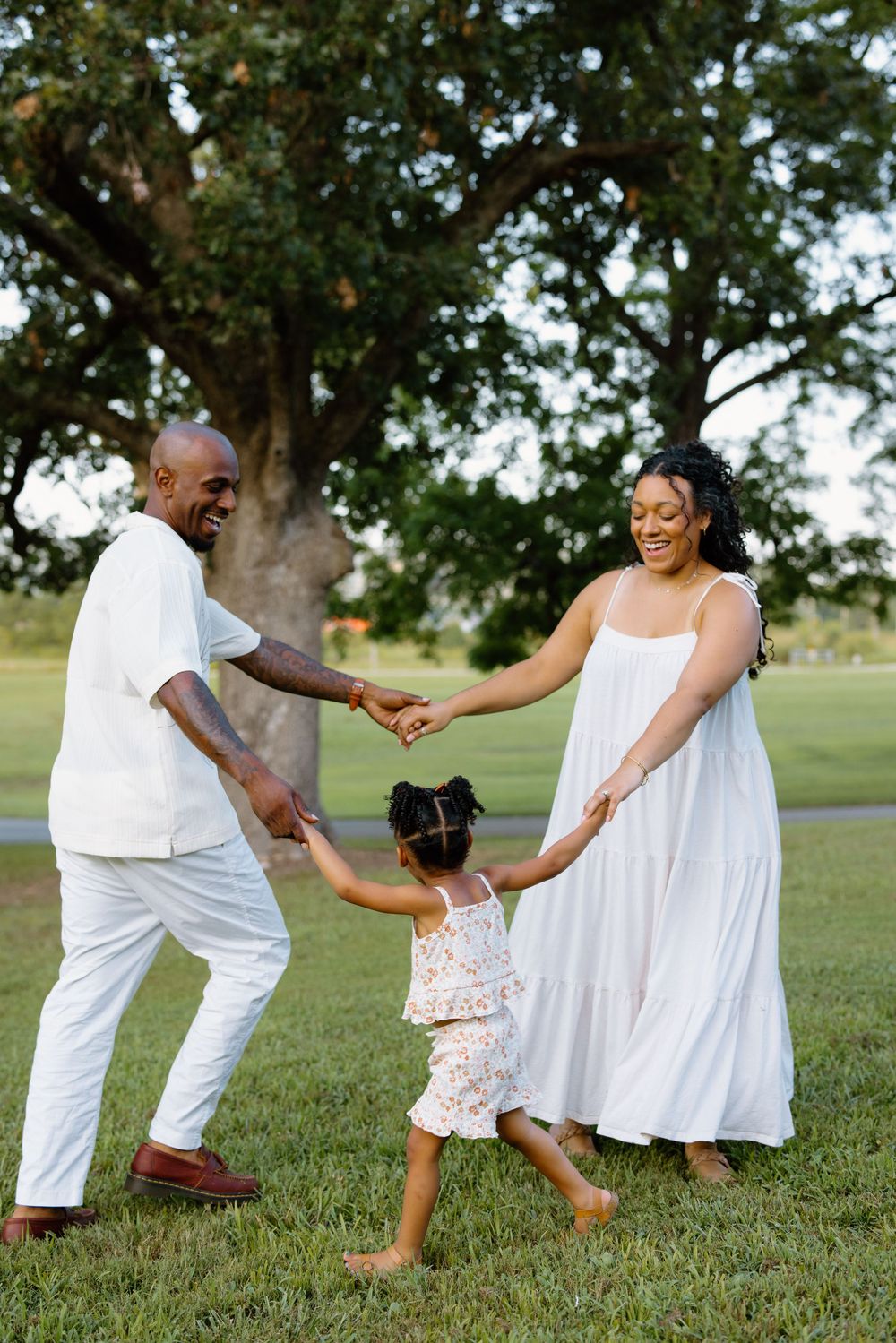 Playful family holding hands and spinning in a circle on grass while wearing white outfits.