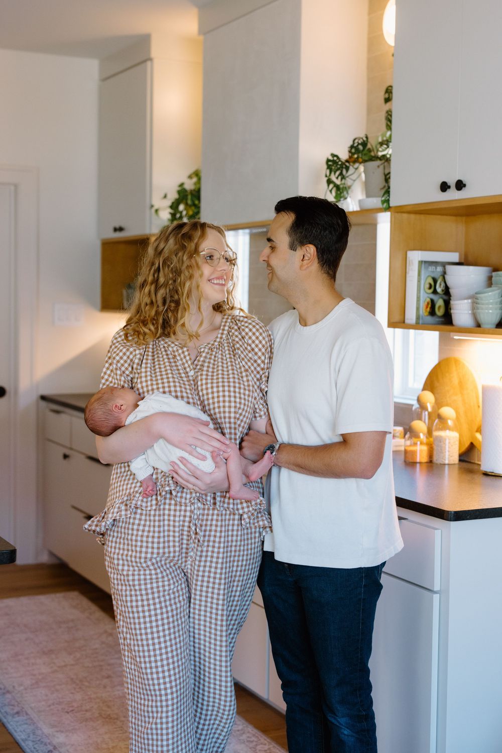 A couple in a modern kitchen space shares a tender moment while holding a baby in pink.