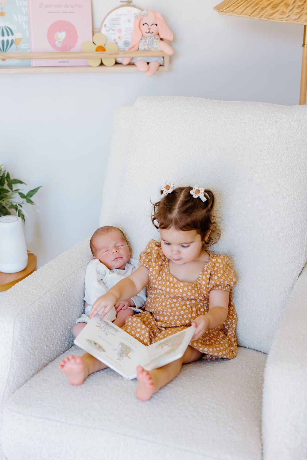 A small child and baby sit together on a white couch while reading a book in matching polka dot outfits.