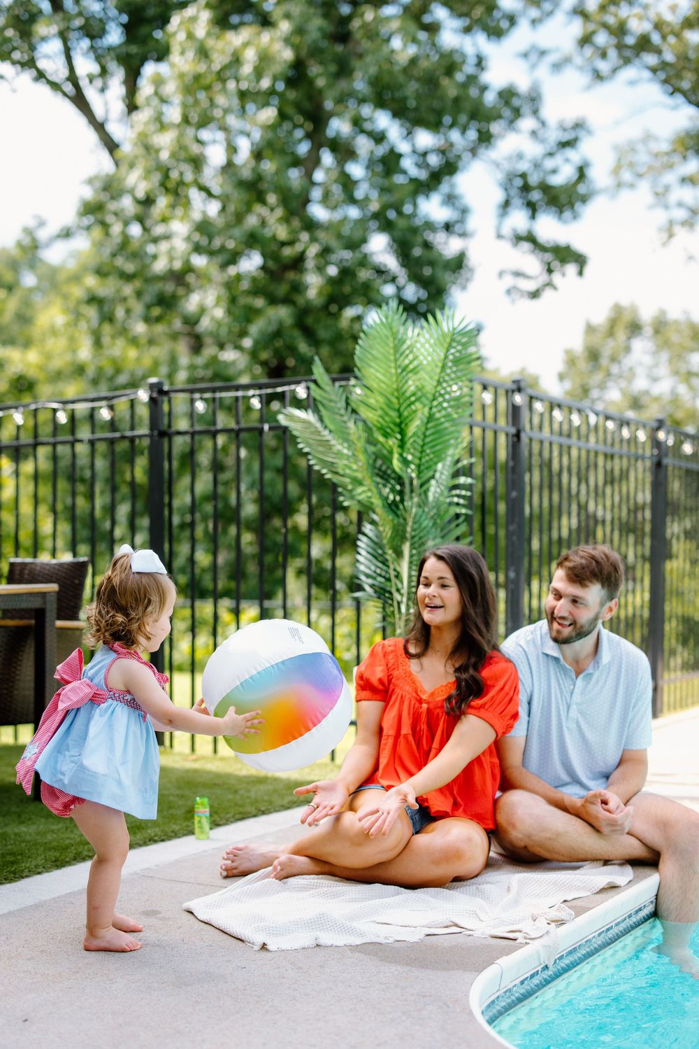 Family enjoying summer by the pool with a toddler playing with a colorful beach ball on a sunny day.