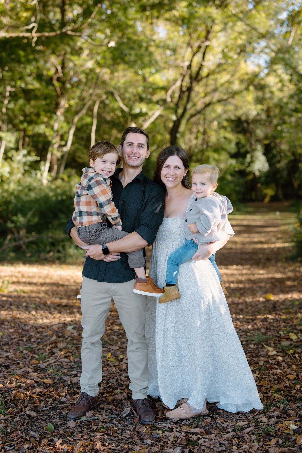 Happy family of four posing outdoors on a leaf-covered path surrounded by lush green trees in autumn.