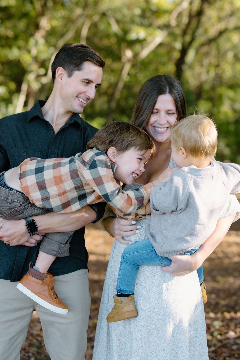 A joyful family of four laughs together outdoors, with parents holding two young boys in a sunlit forest setting.