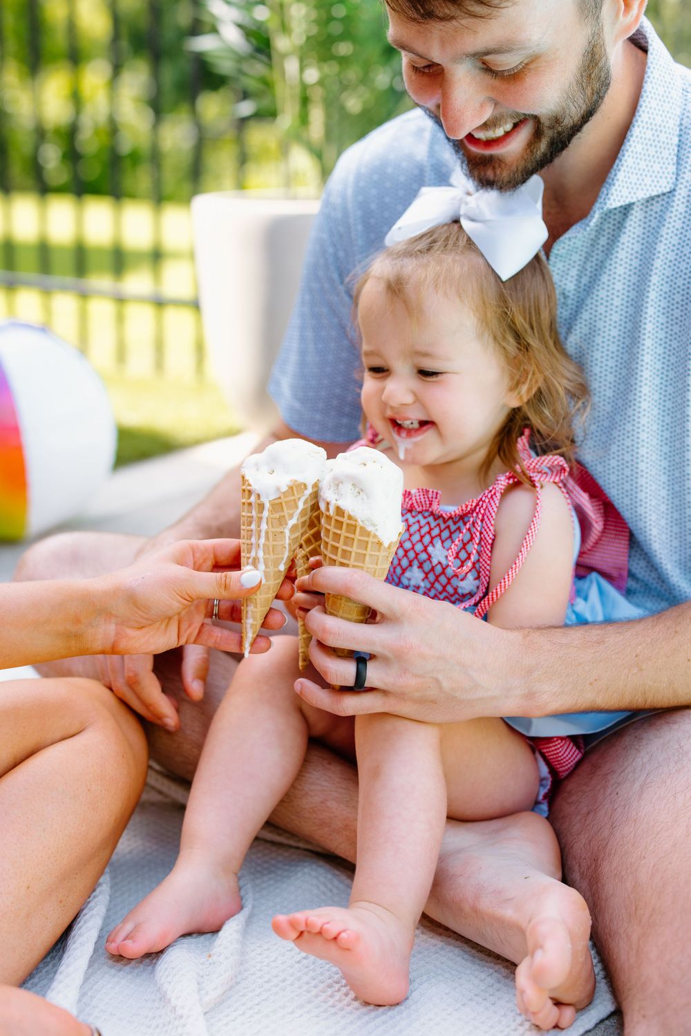 A smiling baby girl sits on her father's lap outdoors, happily holding ice cream cones on a sunny day.