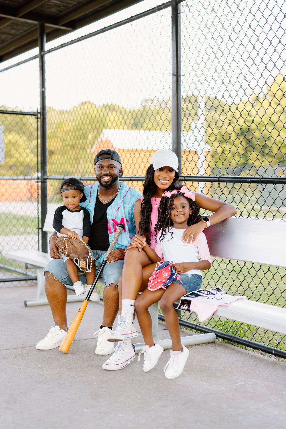 A happy Black family of four poses in a baseball dugout with a bat and gloves, smiling at the camera on a sunny day.