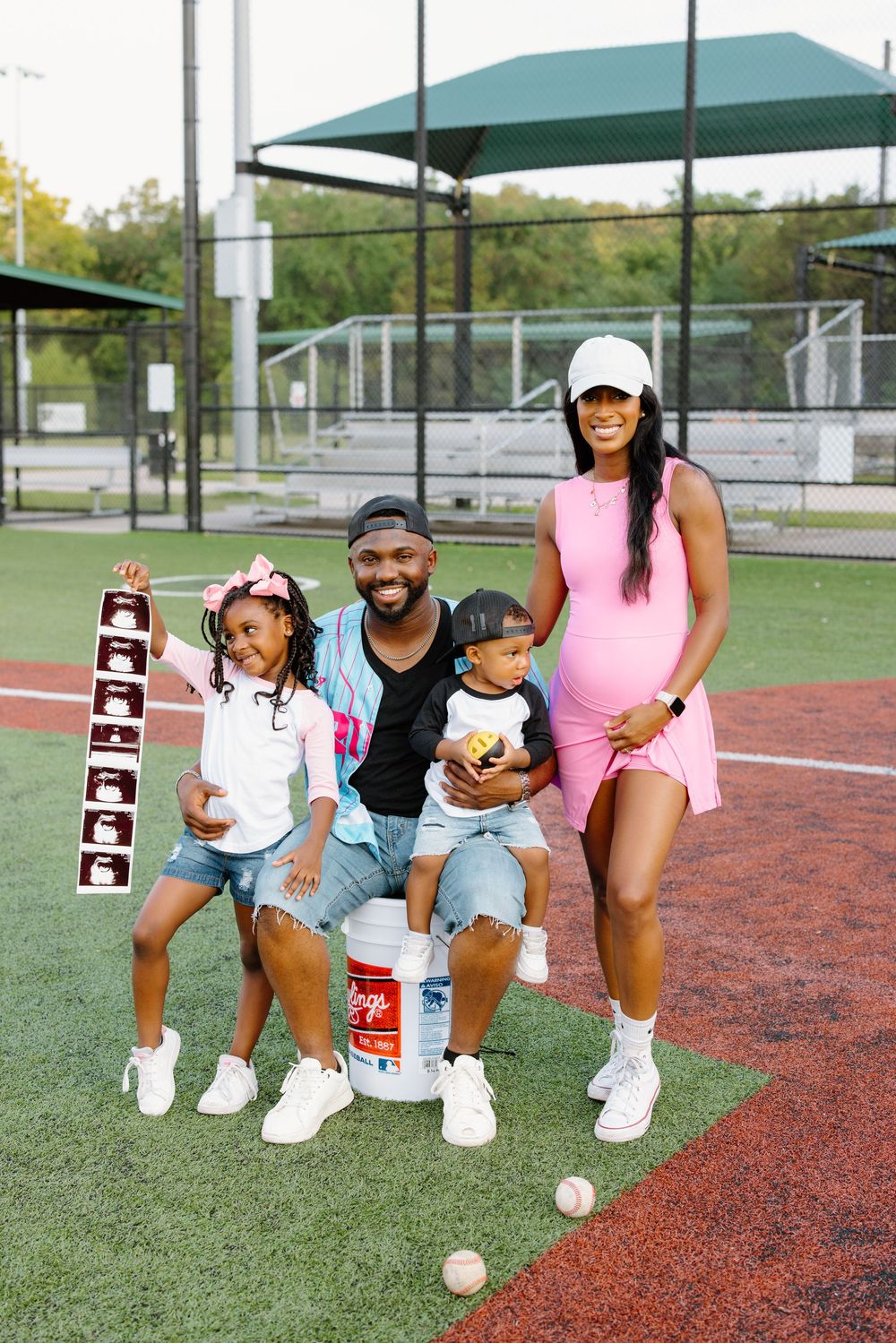 Happy family on baseball field, pregnant mom in pink dress, dad with kids holding ultrasound photos for gender reveal.