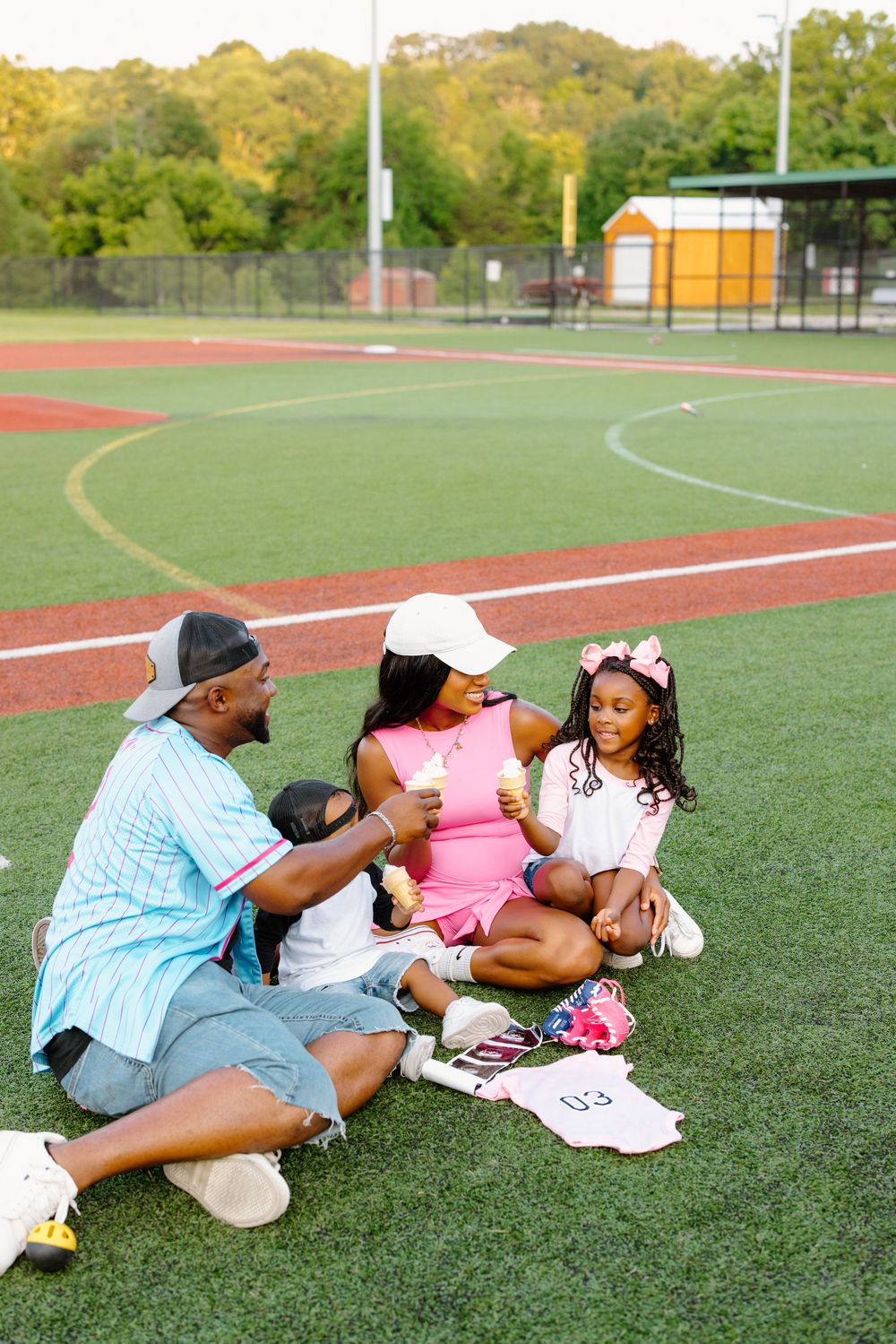 A family sits on a green sports field, sharing ice cream treats on a warm sunny day.