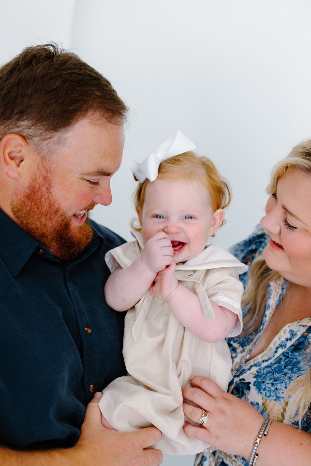 Happy baby girl with white bow laughs while being held by smiling parents in a bright family portrait.