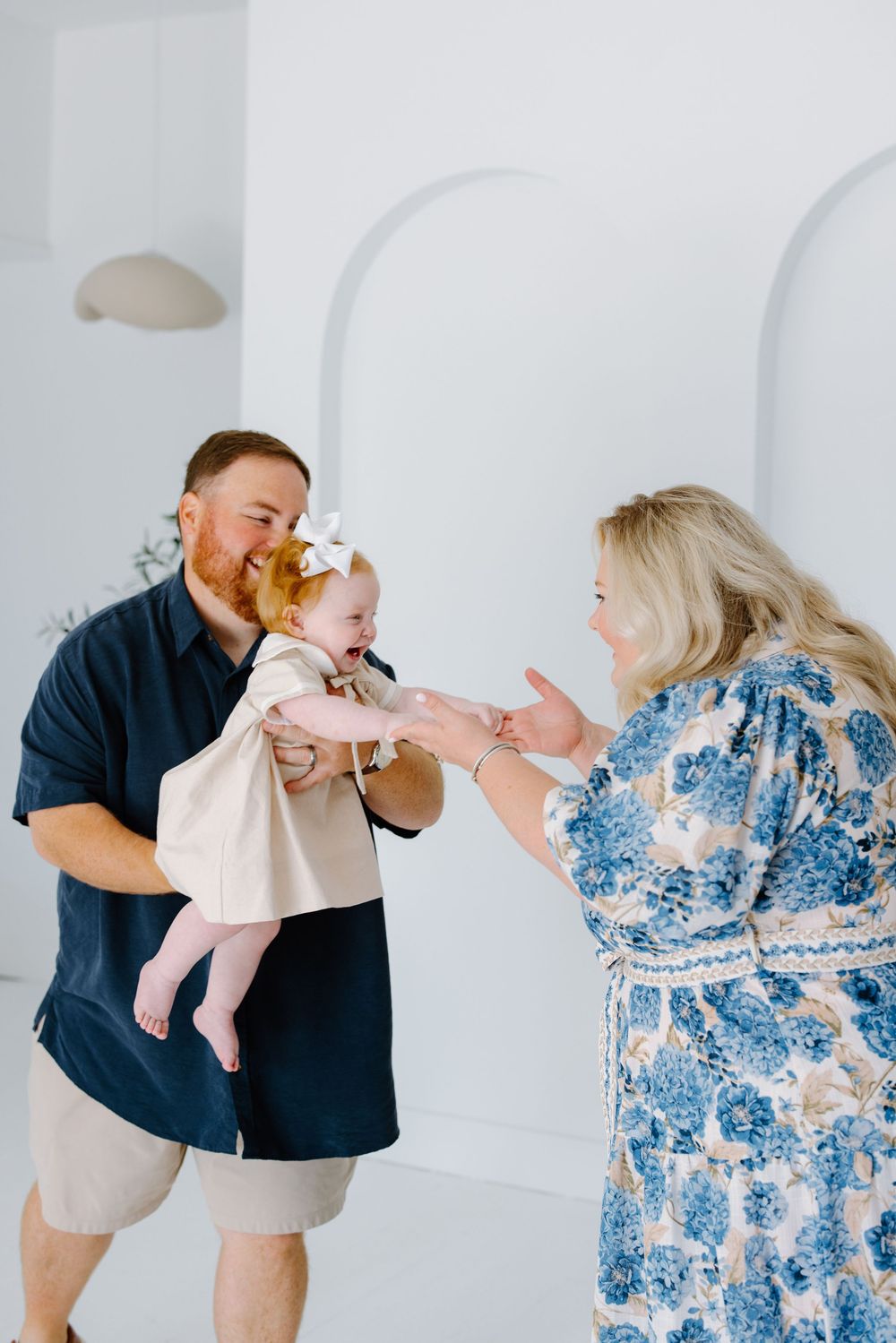 A man holds a smiling baby girl toward a woman in a blue floral dress in a bright white studio setting.