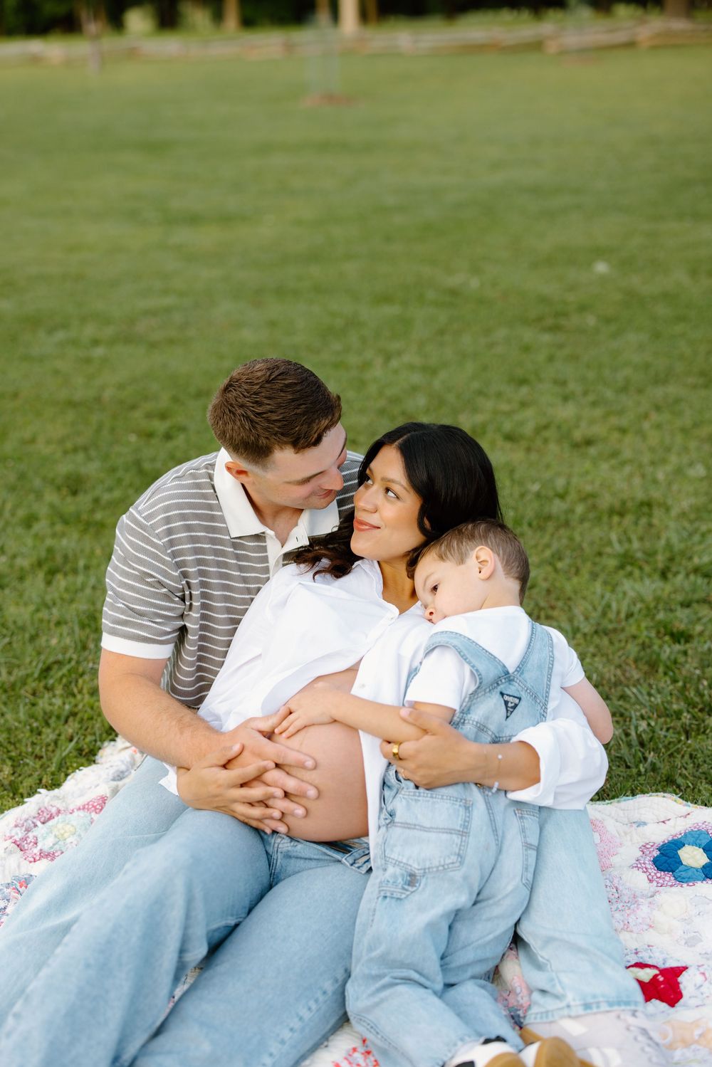 Pregnant woman sits on quilt with husband and toddler in denim overalls on green grass during maternity photo session.