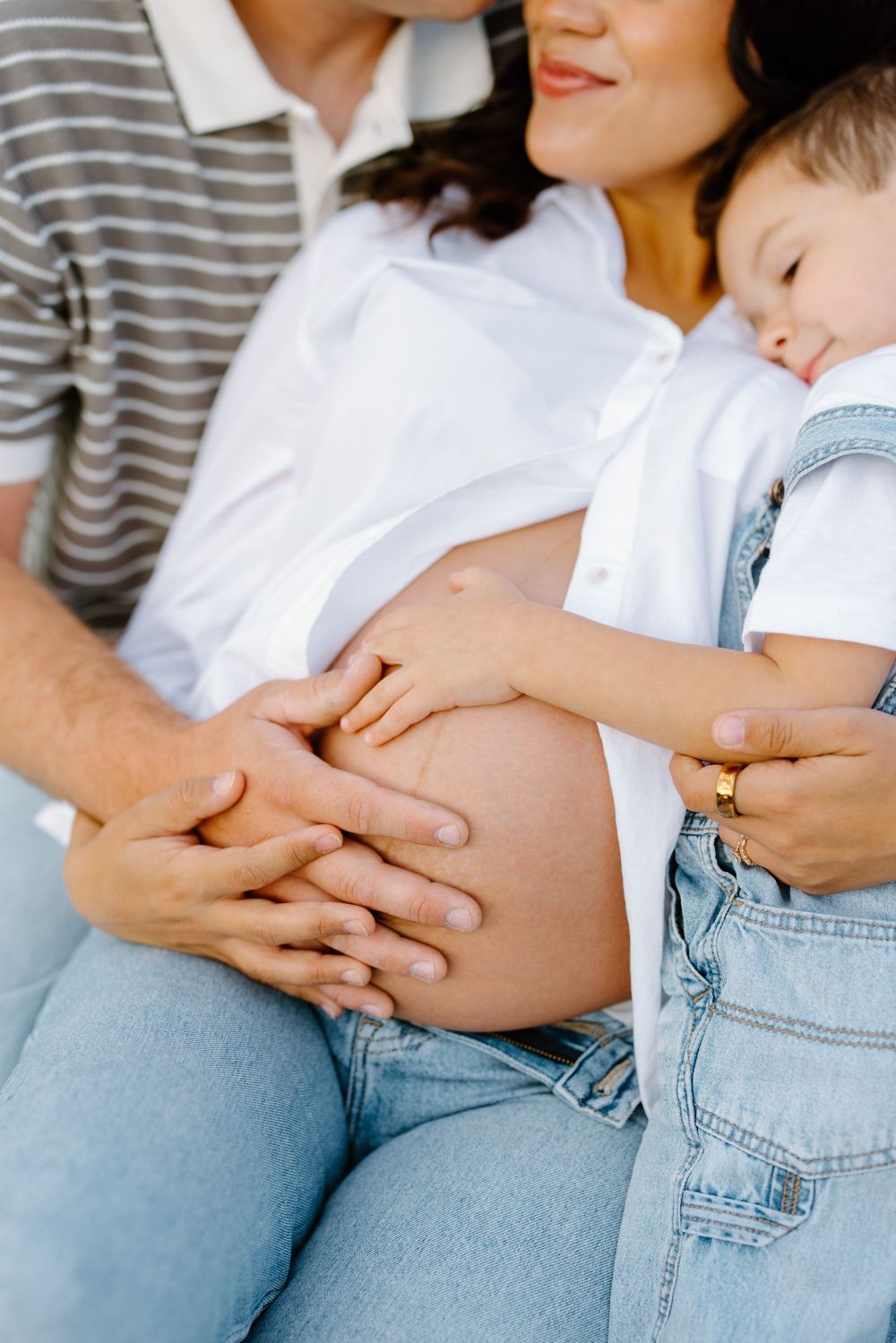Pregnant woman in white shirt and jeans with family hands touching her baby bump, toddler resting on her shoulder.