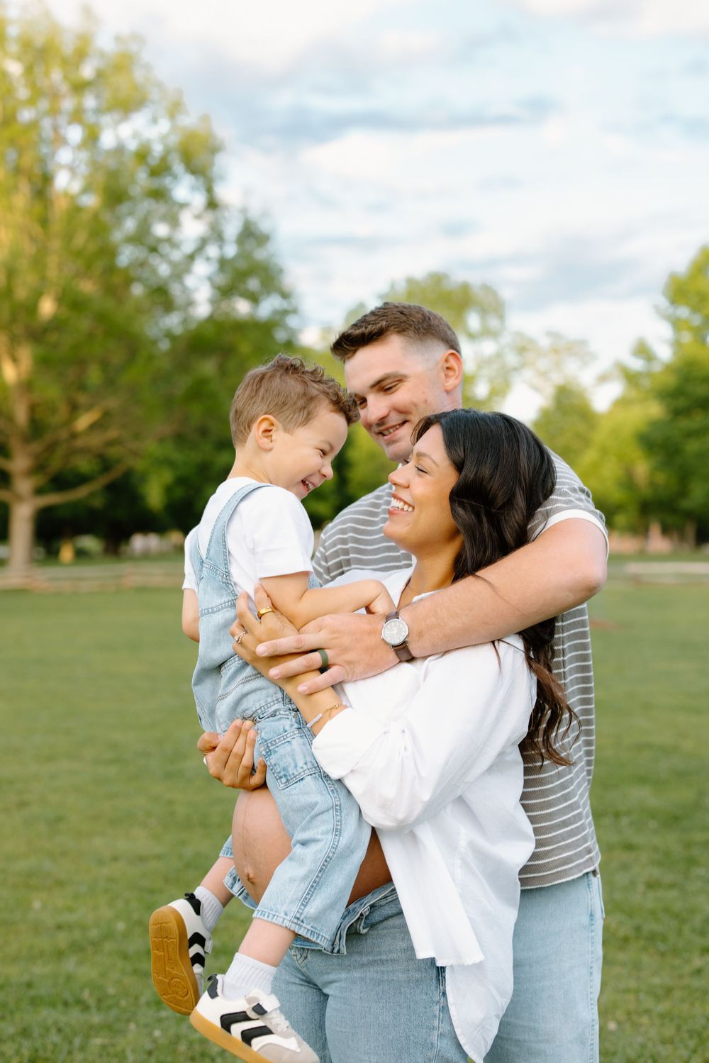 Happy young family with toddler boy laughing together in a sunny green park during a warm summer day.