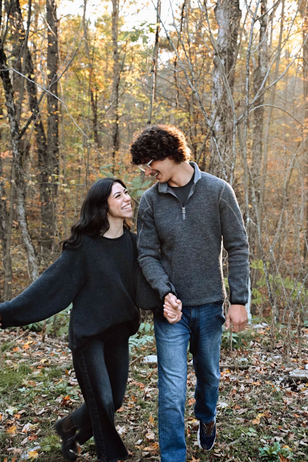 A couple holds hands while walking through an autumn forest with golden leaves and bare trees in the background.