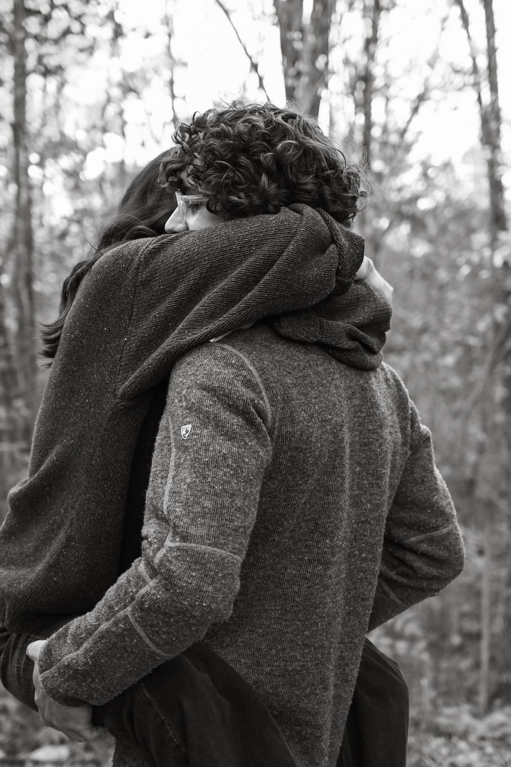A black and white photograph of a couple embracing in a wooded forest setting.