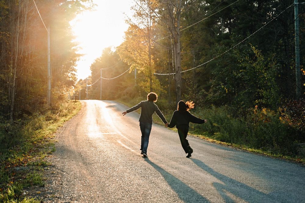 Two people joyfully running down a sunlit forest road during golden hour as sunbeams filter through autumn trees.