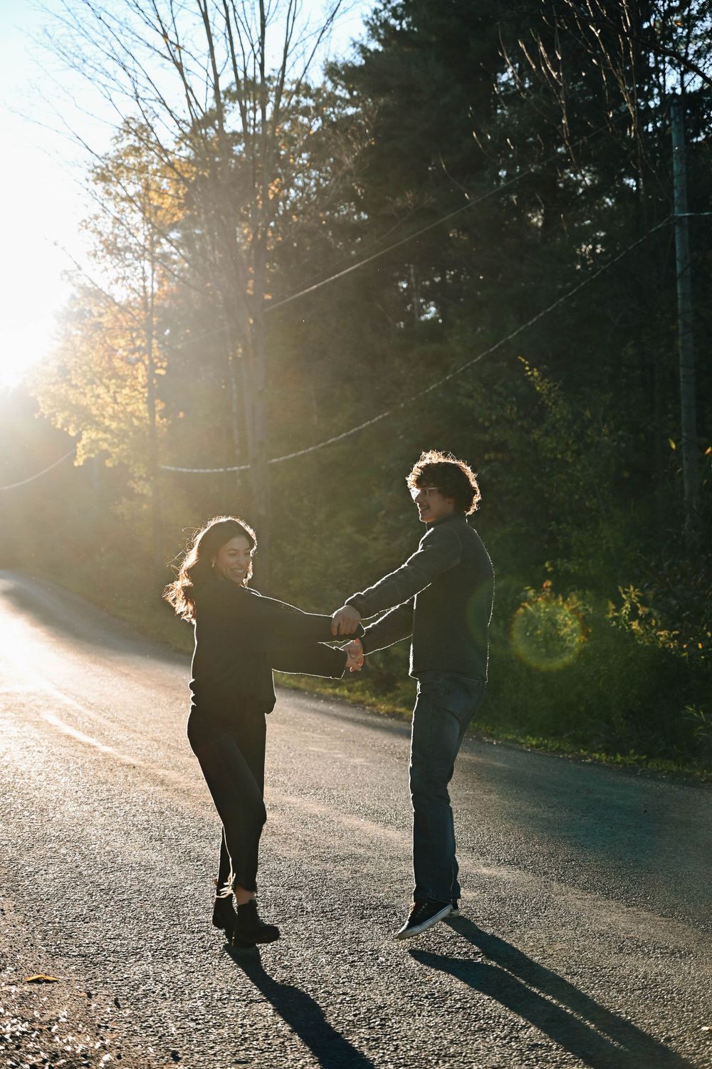 Two people holding hands and standing together on a sunlit road with trees and lens flare in the background.