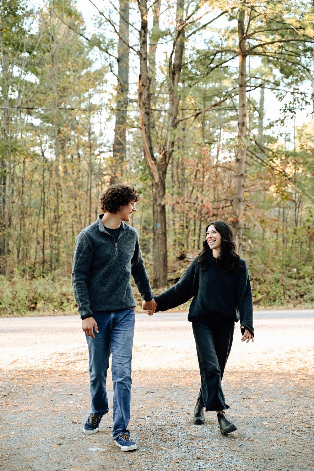 A couple holds hands while walking together on a wooded autumn path wearing casual sweaters and jeans.