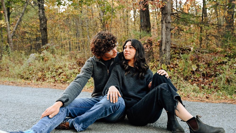 A couple sits together on a road surrounded by autumn trees, sharing an intimate moment in casual attire.