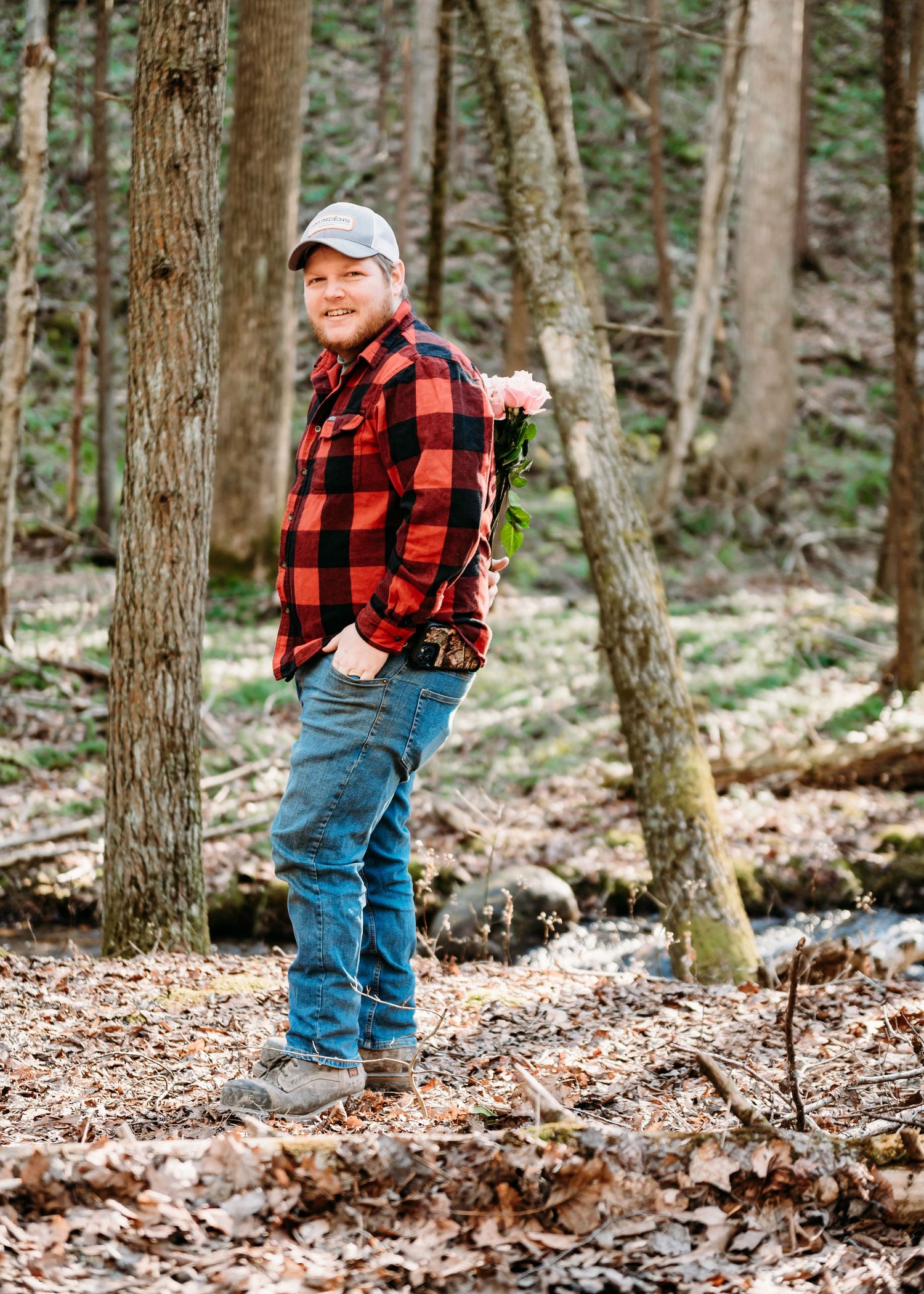 A person in a red buffalo plaid shirt and baseball cap poses in a wooded forest setting.