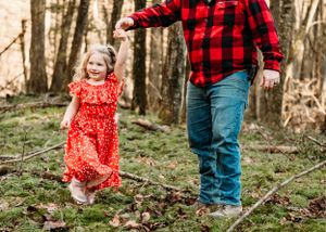 A playful moment of father and daughter in the forest featuring matching red outfits and joyful dancing among fallen leaves.