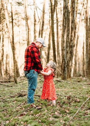 A heartwarming woodland scene with a father in red flannel shirt and daughter in bright red polka dot dress against a backdrop of trees.