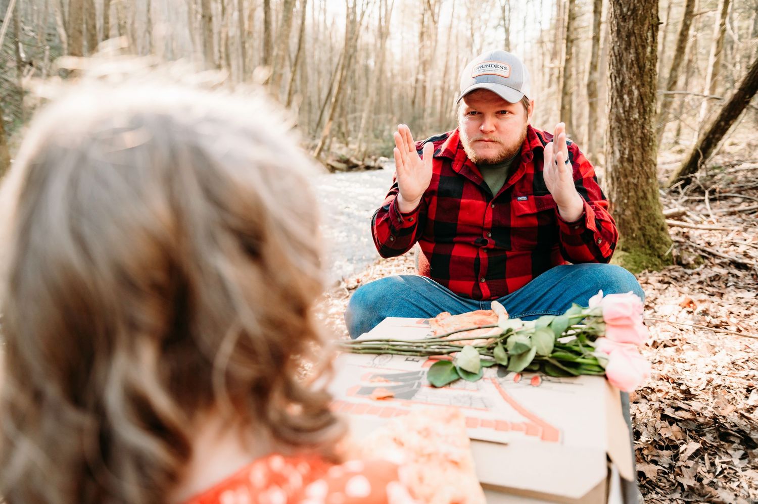 Person in red plaid shirt sits on blanket in forest setting during outdoor gathering.