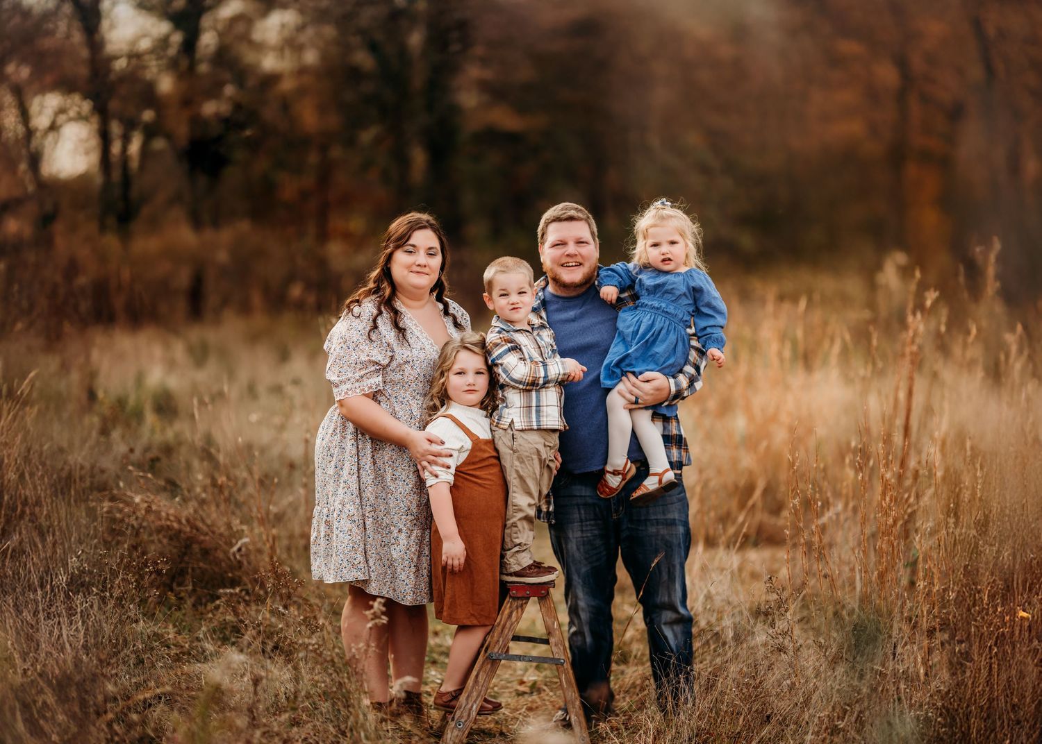 A family stands together in a rustic autumn field with tall brown grass and trees in the background during golden hour.