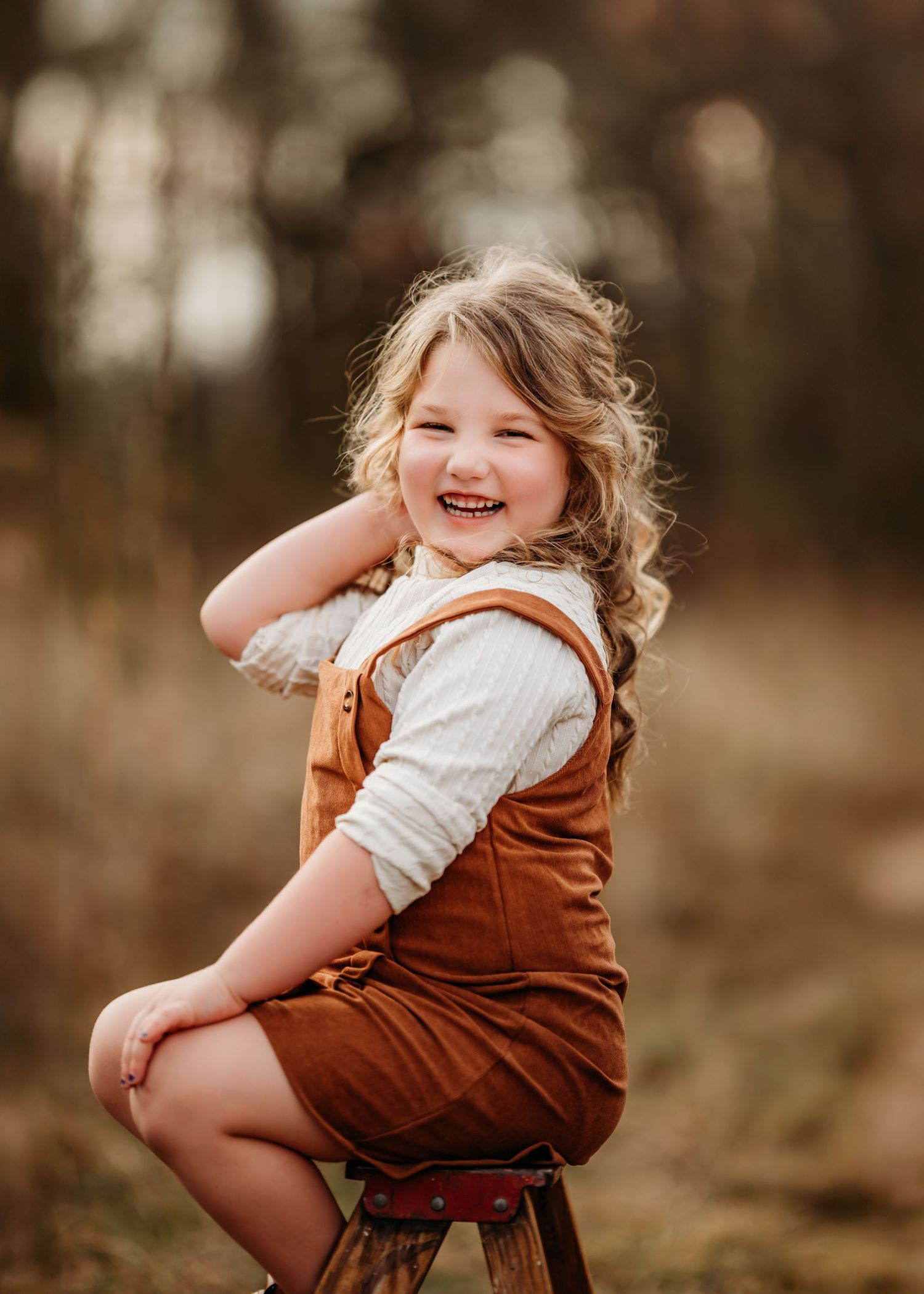 Child in brown corduroy overalls and white sweater sits on wooden stool during outdoor autumn photoshoot.