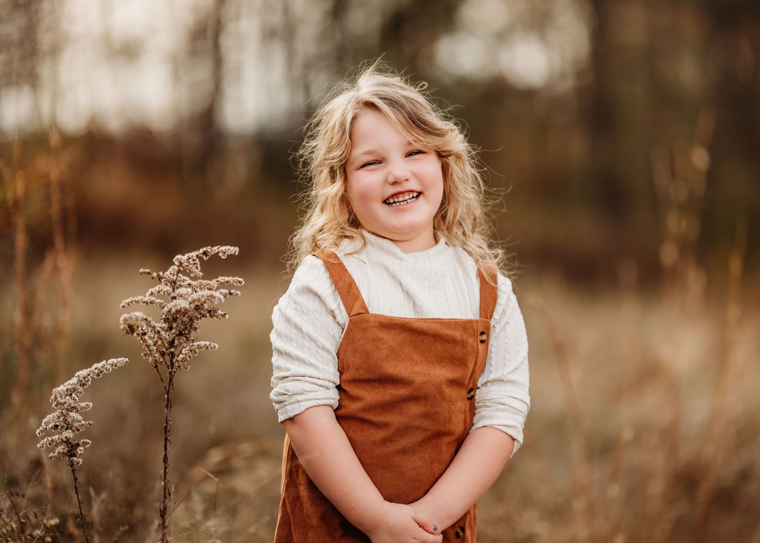 Child in brown corduroy overalls and white shirt smiling in an autumn field with dried wildflowers.