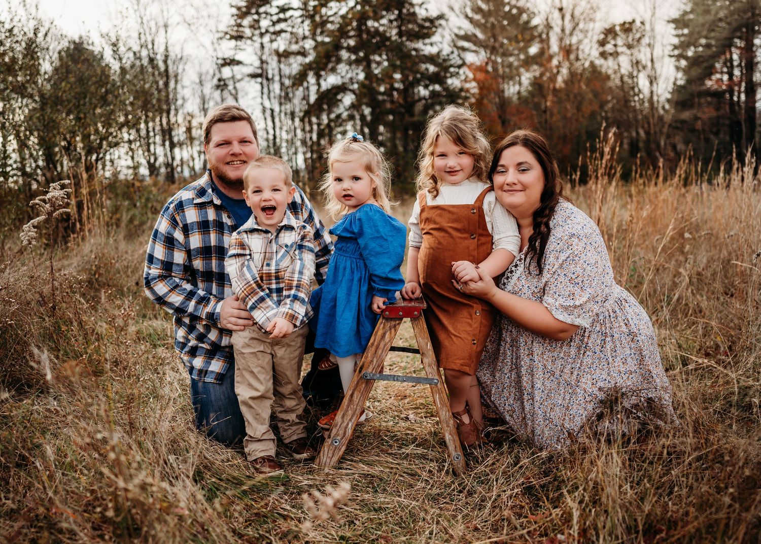 A family poses together in a rustic outdoor setting with tall grass and autumn trees in the background.