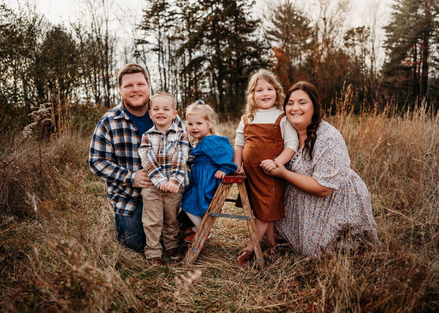 A family sits together in a rustic outdoor setting with tall grass and evergreen trees in the background.