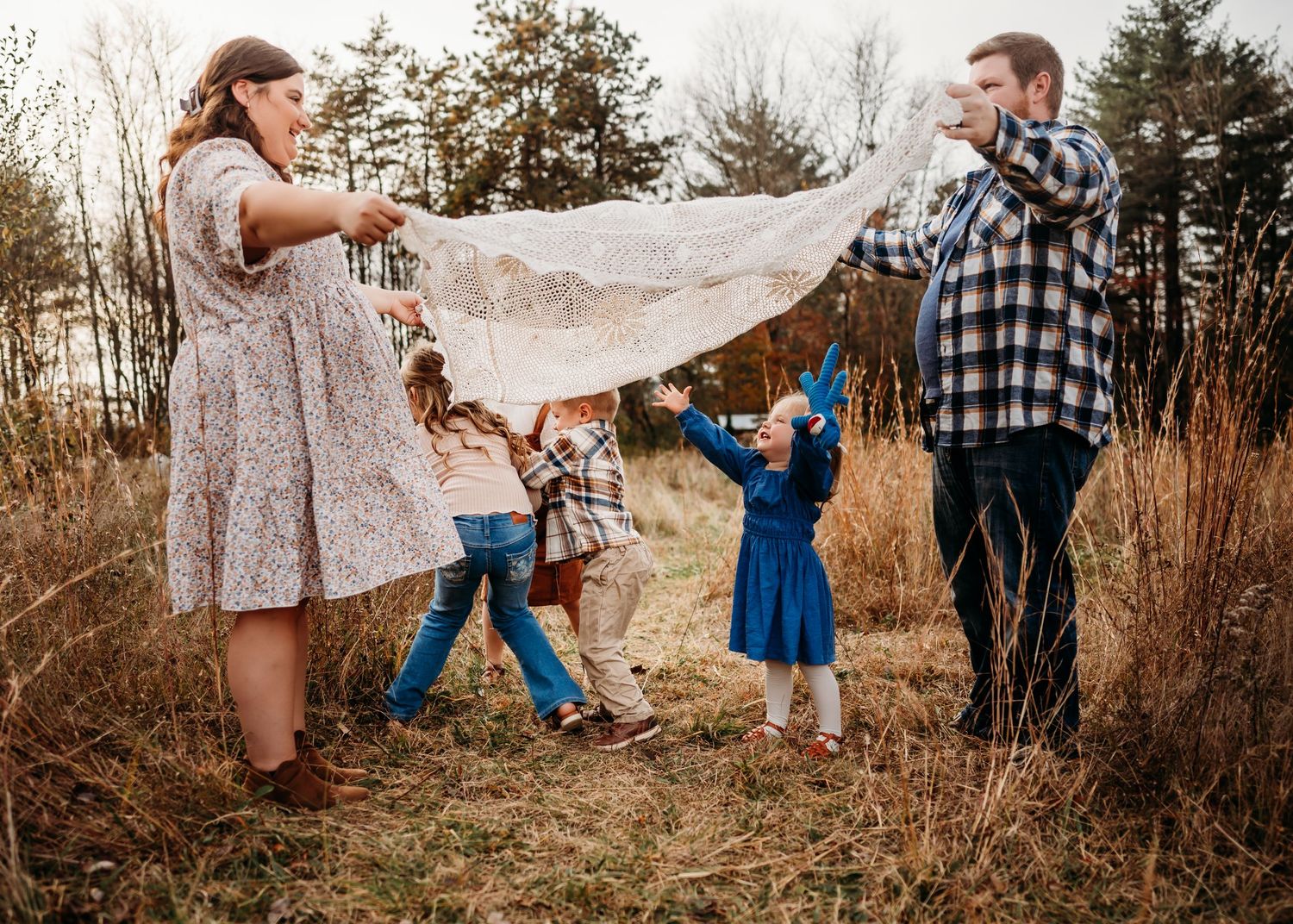 Family plays together holding a blanket in a golden field during sunset while wearing coordinated outfits.
