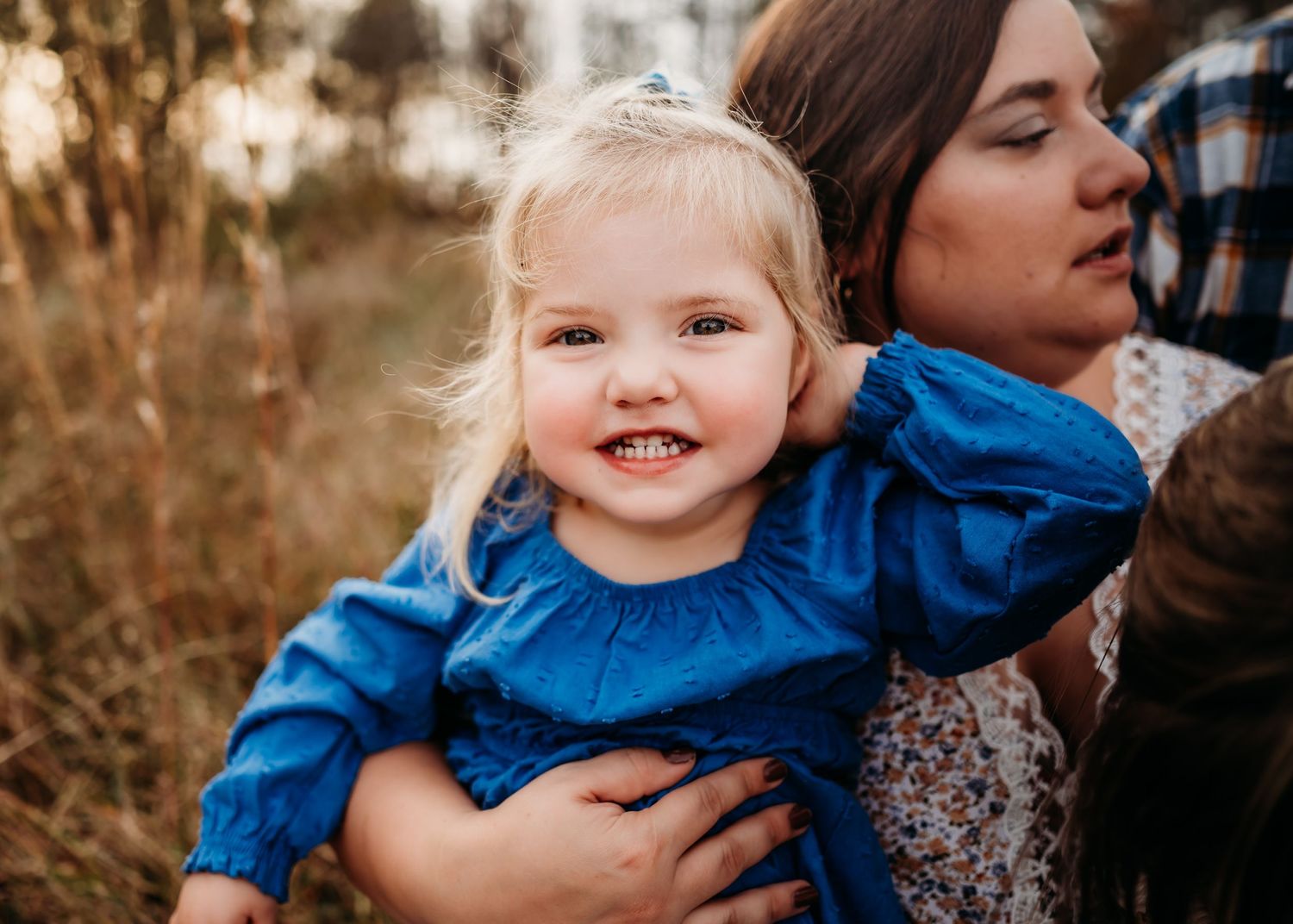 Young child in bright blue top smiles joyfully while being held outdoors during golden hour.