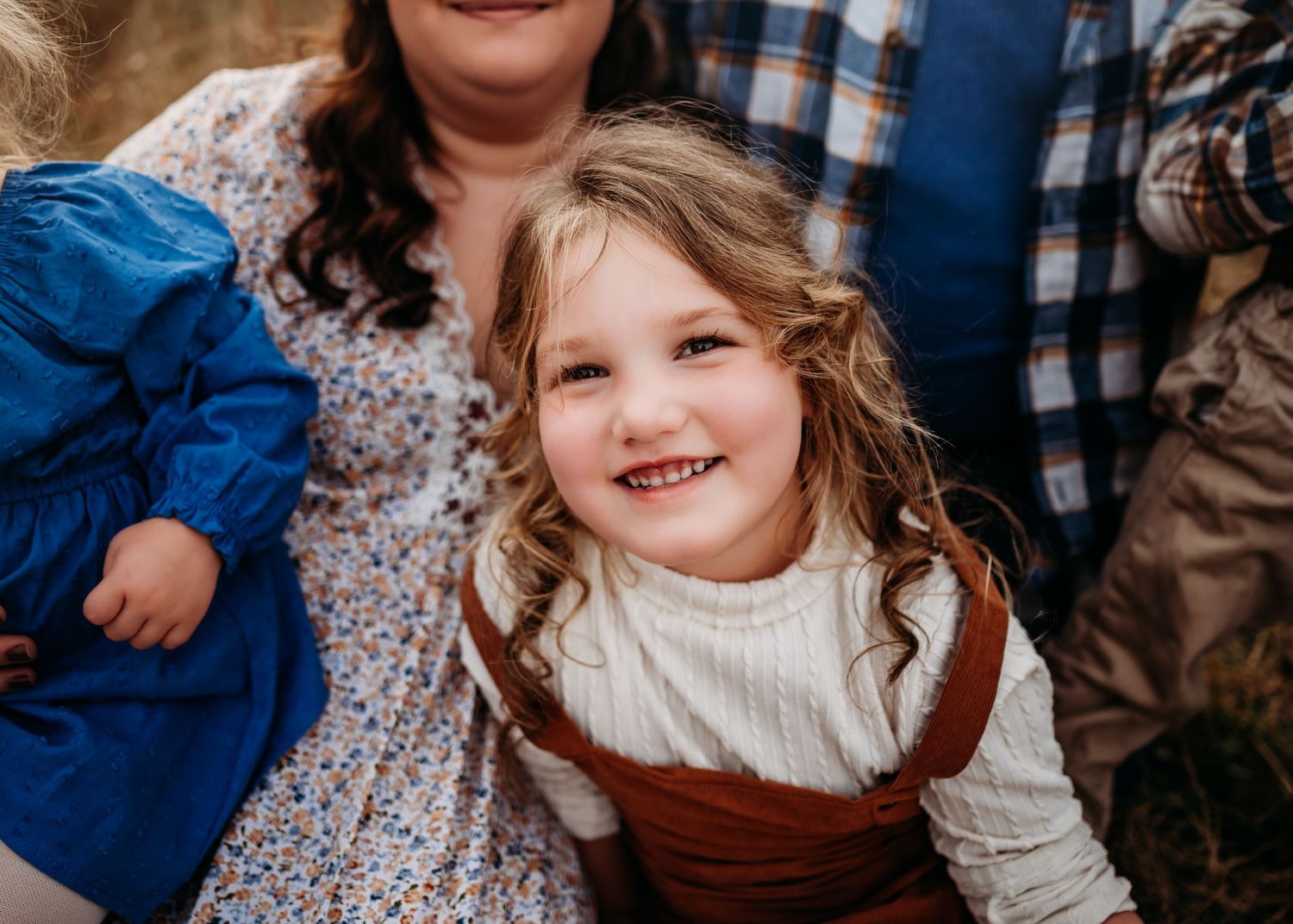 Joyful child wearing a white sweater and brown overalls smiles brightly during a family photo session.