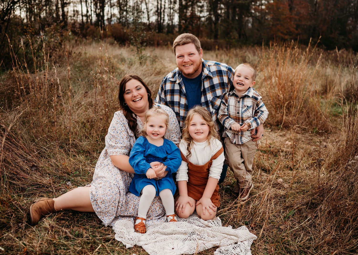 A family sits outdoors on a blanket in a field of tall grass during autumn while wearing coordinated outfits.