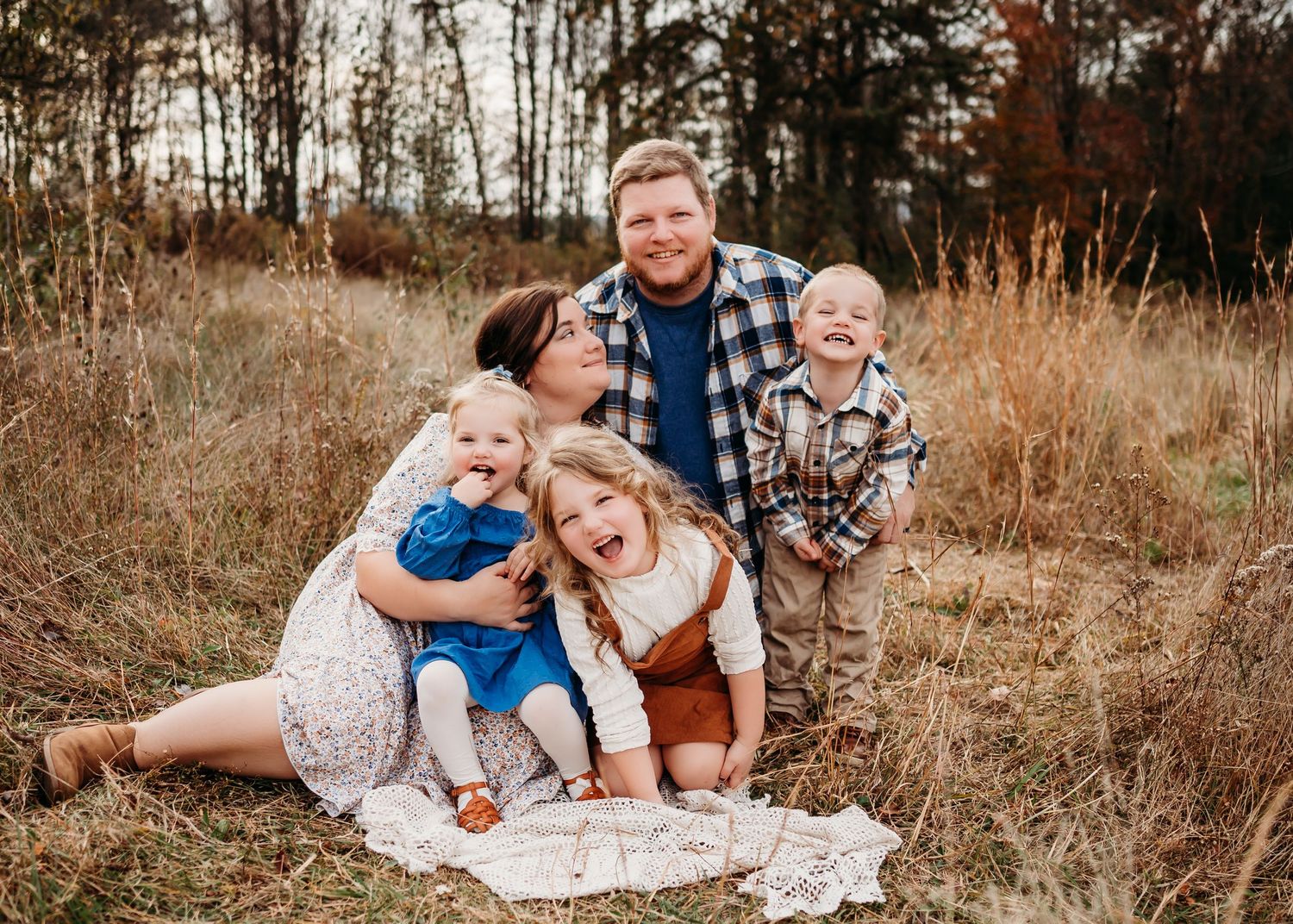 Family of four sitting together on blanket in autumn field with tall grass and trees in background.