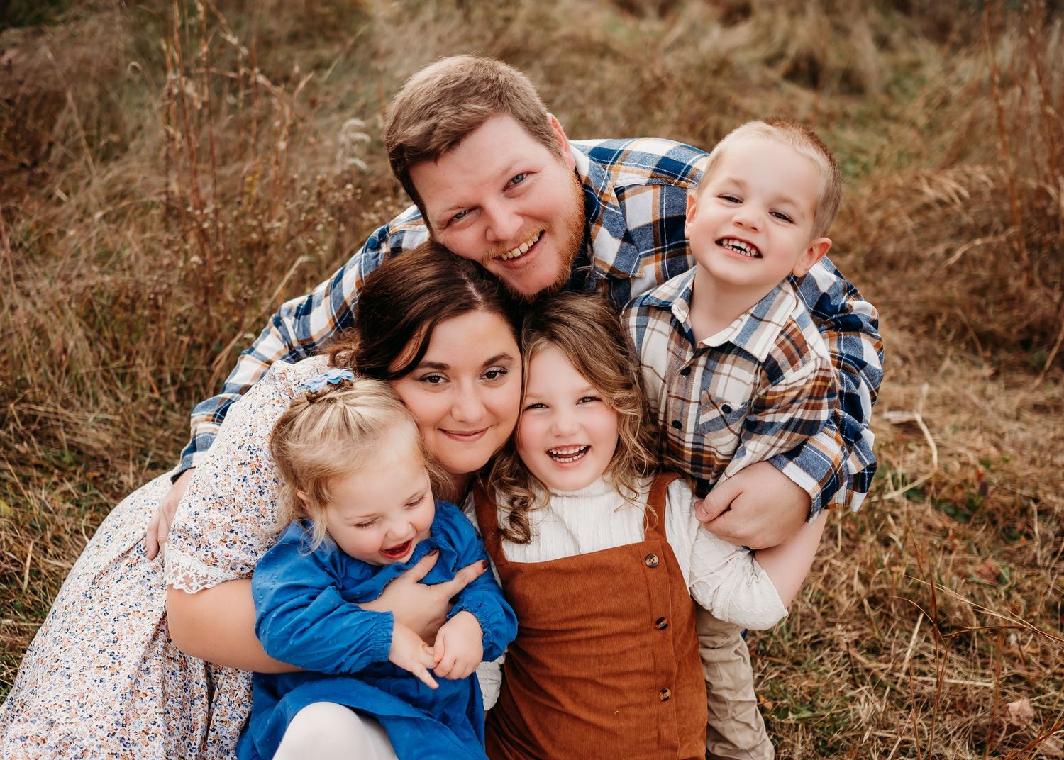 A family of five snuggled close together and smiling while sitting in a natural outdoor setting with brown grass.