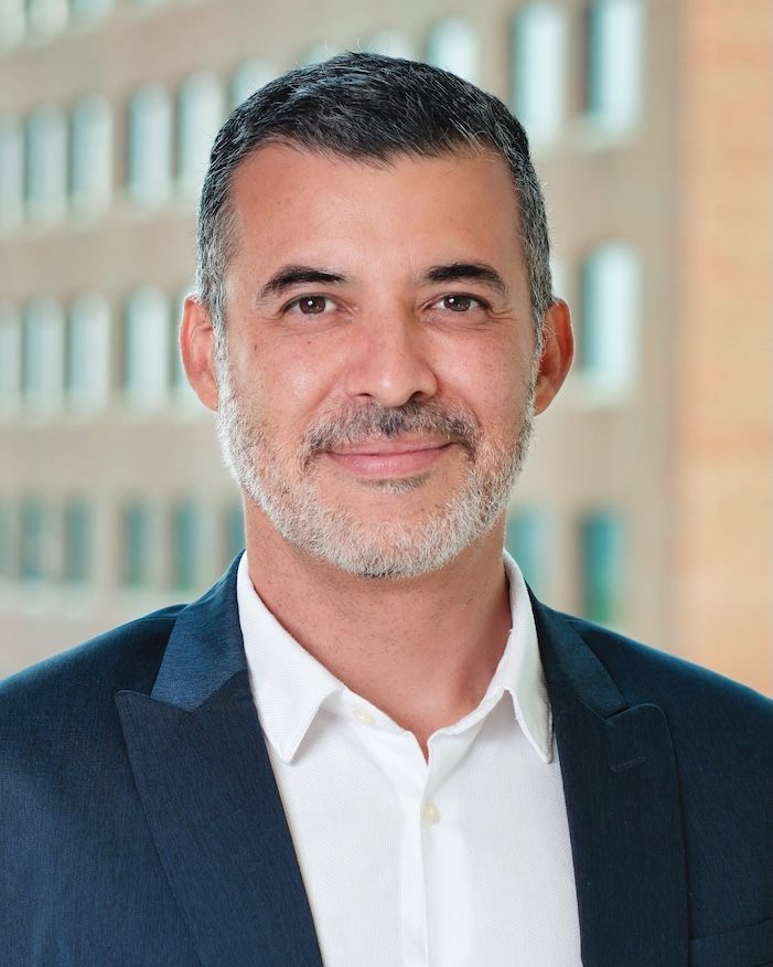 Professional headshot of a man with salt and pepper beard wearing a navy suit against an office building.