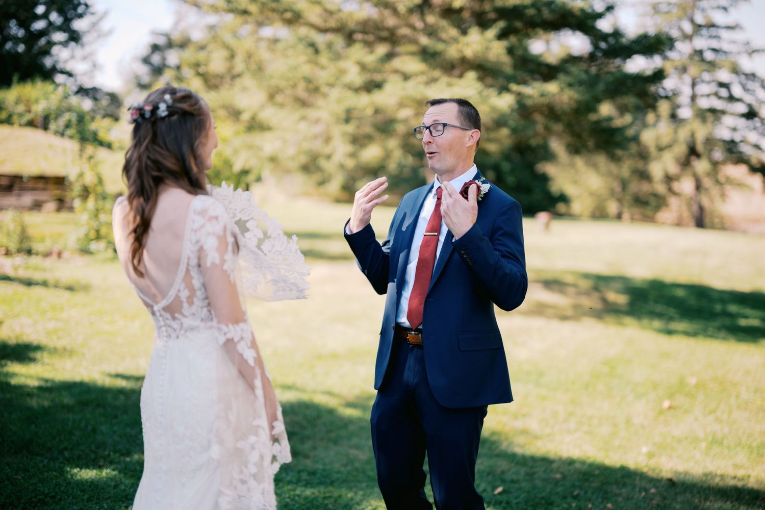 Two people share an emotional moment outdoors on a sunny day, with one wearing a lace dress and another in a navy suit.