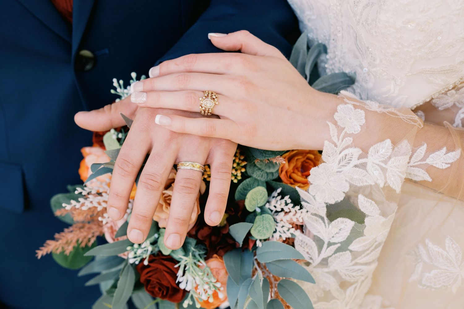 Hands showing wedding rings resting on autumn-colored bouquet with eucalyptus and rust-colored flowers.
