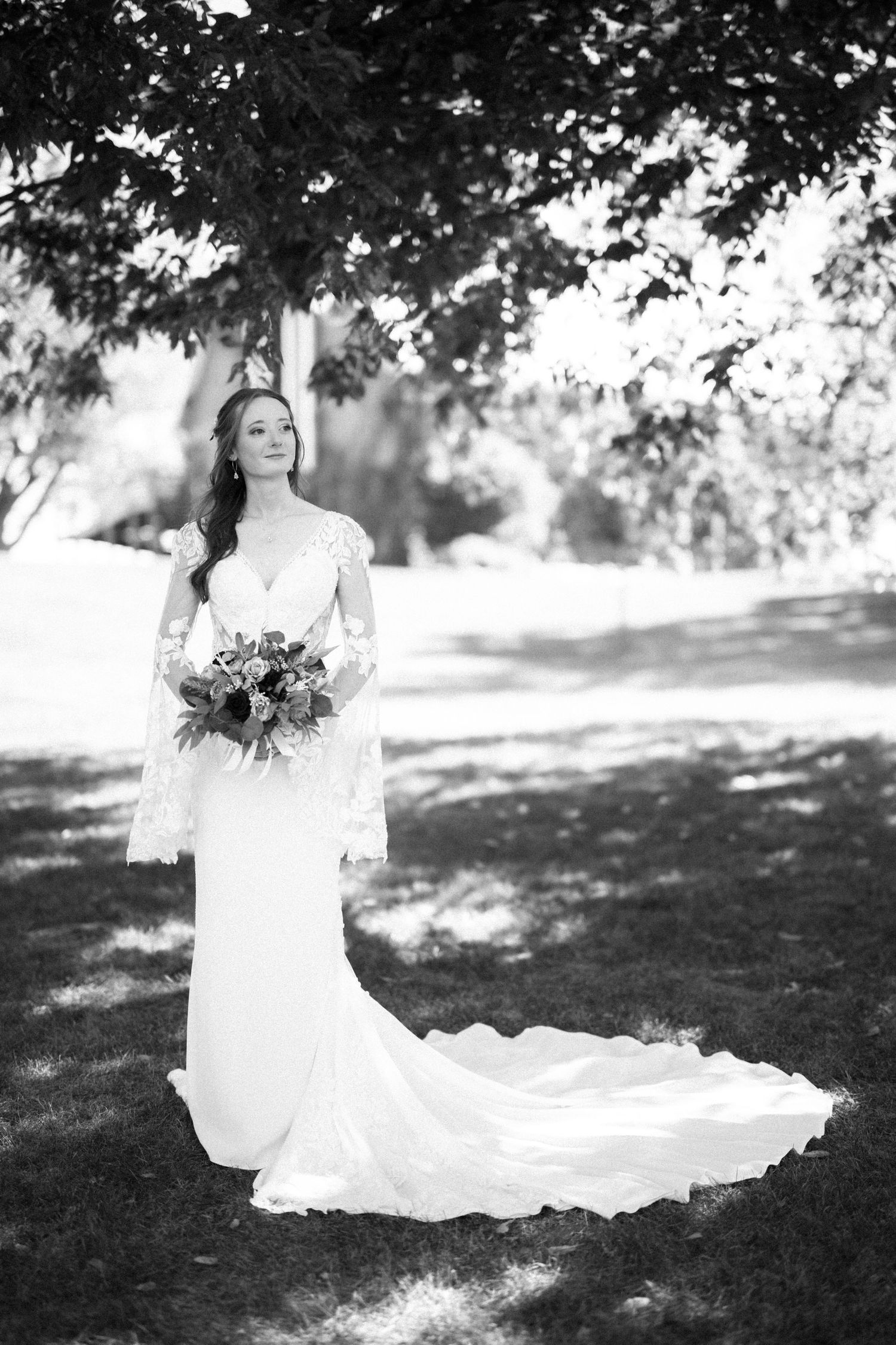 Black and white portrait of a bride in a lace wedding gown with long train holding a bouquet in a garden setting.