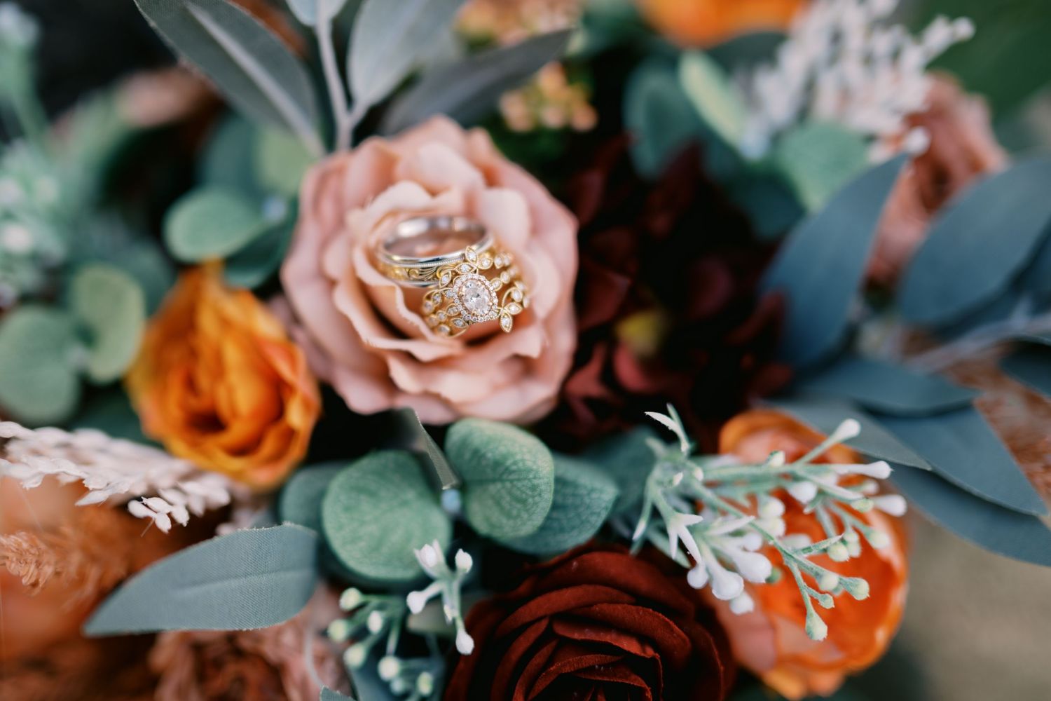 Wedding rings rest atop a dusty pink rose surrounded by orange blooms and sage green foliage in a romantic bouquet.