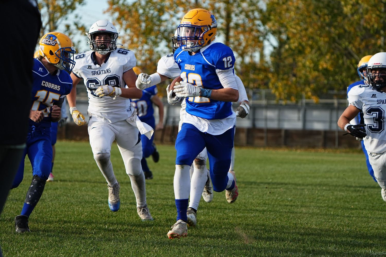 High school football player in blue and gold uniform runs with the ball during game while defenders give chase.