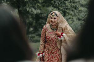 A bride in an ornate red and gold traditional lehenga poses during an outdoor wedding ceremony.