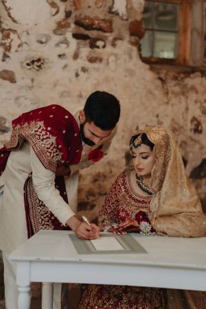 A traditional South Asian wedding couple signing their marriage certificate at a rustic venue.