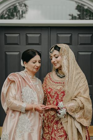 Two women in traditional South Asian attire share an intimate moment looking at jewelry by a dark doorway.