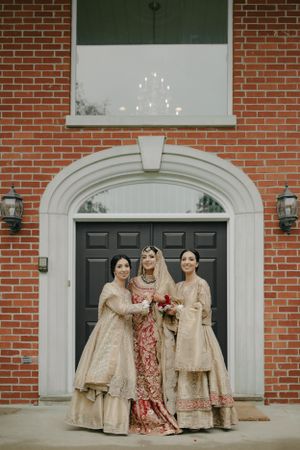 Three people in traditional South Asian wedding attire pose in front of a brick building with an arched doorway.