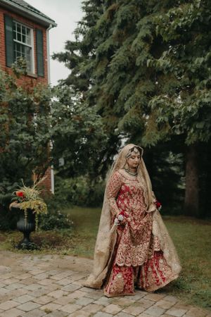 South Asian bride in red and gold lehenga standing in garden setting near brick building.