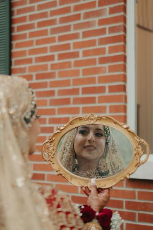 Reflection of bride wearing ornate jewelry and veil in decorative golden mirror against brick wall.
